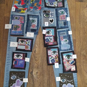 Pode incluir: Jeans de ganga azul claro adornados com vários patches de tecido. Os patches apresentam desenhos de olhos, padrões florais e formas abstratas numa mistura de cores, incluindo azul, verde, vermelho e rosa. Os jeans são exibidos numa superfície de madeira.