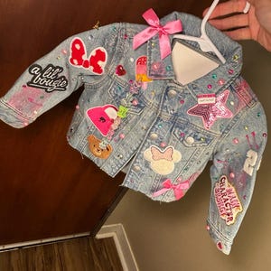 May include: A light blue denim jacket adorned with patches, rhinestones, and a pink bow. Patches include a Minnie Mouse bow, a teddy bear, and the text "a lil bougie". The jacket is embellished with colorful rhinestones and sequins.