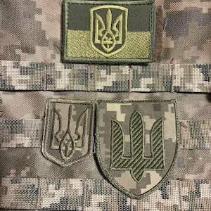 Peut inclure: Trois écussons brodés représentant les armoiries ukrainiennes, le Tryzub, dans des tons de vert et de beige. Les écussons sont rectangulaires et en forme de bouclier, avec un motif de camouflage.