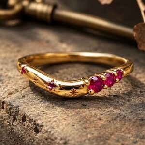 May include: A gold curved ring featuring three prominent round red rubies set in a cluster, accented by smaller embedded rubies and engraved star motifs along the band. The polished metal surface reflects light, highlighting the intricate star details.