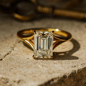 Op de afbeelding: Een gouden verlovingsring met een smaragdgeslepen diamant. De ring heeft een split-schacht ontwerp. De diamant is gezet in een vier-klauwzetting. De ring ligt op een stenen oppervlak.