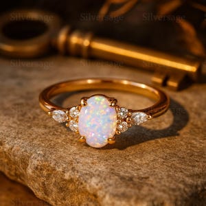 Könnte beinhalten: Ein goldener Ring mit einem ovalen Opal-Mittelstein und kleinen Diamantakzenten. Der Opal ist von einem Kranz kleinerer Diamanten umgeben und auf einem goldenen Band gefasst. Der Ring wird auf einer strukturierten, braunen Oberfläche präsentiert.