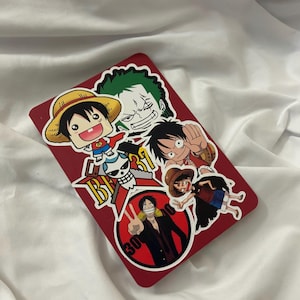 May include: A red rectangular item covered in colorful stickers featuring anime characters. The stickers depict various characters in action poses, with bold outlines and vibrant colors. The background is a white, wrinkled fabric.