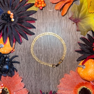 May include: A gold-colored bracelet with a textured surface, set against a wooden background. The bracelet is surrounded by fall-themed decorations, including orange and black flowers, spiders, and pumpkins.