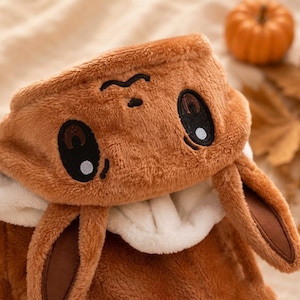 May include: A brown plush hooded garment featuring an embroidered face with large dark eyes and a small nose. The hood includes two long, soft, brown ears with dark brown inner accents and a white interior lining, crafted from a fuzzy fabric.
