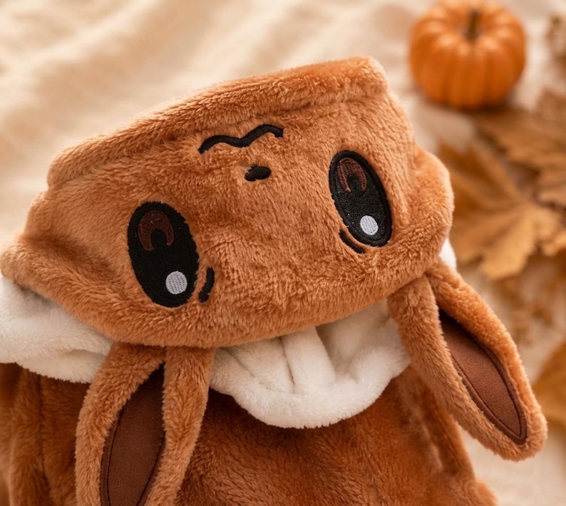 May include: A brown plush hooded garment featuring an embroidered face with large dark eyes and a small nose. The hood includes two long, soft, brown ears with dark brown inner accents and a white interior lining, crafted from a fuzzy fabric.