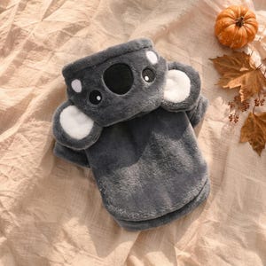 May include: A dark gray plush sweater featuring a koala face design on the hood. The hood includes two large, rounded ears with white centers, a black nose, and two eyes. The garment is made of soft, fuzzy material with long sleeves and a hem.