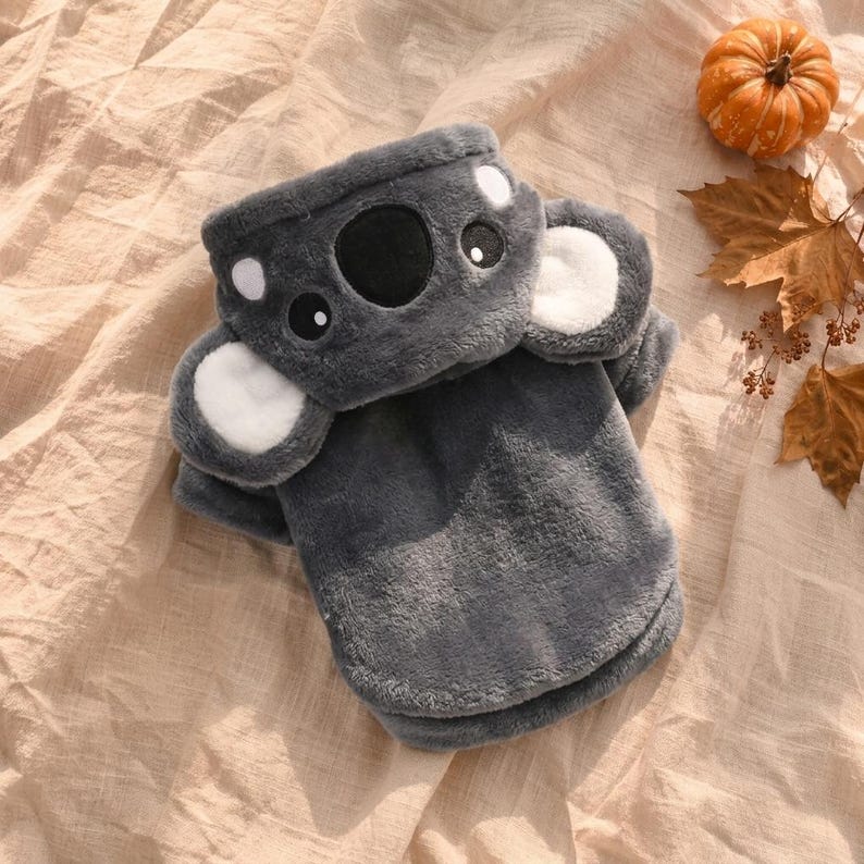 May include: A dark gray plush sweater featuring a koala face design on the hood. The hood includes two large, rounded ears with white centers, a black nose, and two eyes. The garment is made of soft, fuzzy material with long sleeves and a hem.