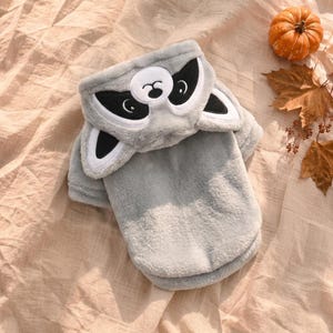 May include: A soft, light gray plush hoodie featuring a raccoon face design on the hood. The hood includes embroidered black and white eye patches, a small black nose, and two protruding ears. The garment is made of a fuzzy, textured fabric.