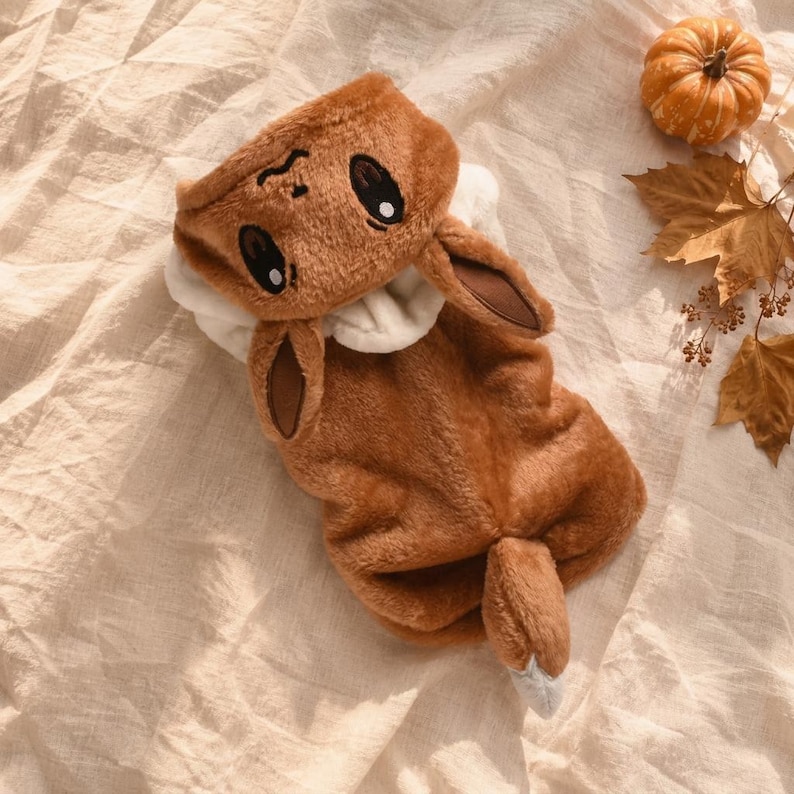 May include: A brown plush hooded garment featuring an embroidered face with large dark eyes and a small nose, two long pointed ears, and a tail with a white tip. The soft, fuzzy material covers the body, and the hood includes a white inner lining.