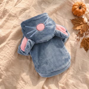 May include: A plush, blue hooded garment featuring a hood designed with a pink nose and embroidered whiskers. The hood includes two soft, pink-lined ears extending from the sides. The fabric has a fuzzy, textured appearance throughout the piece.