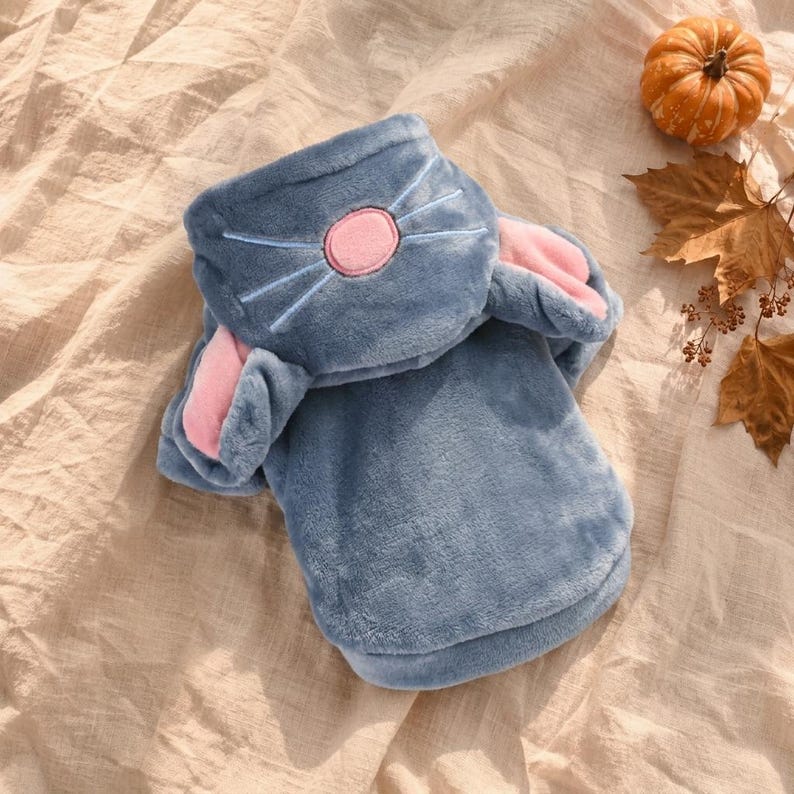 May include: A plush, blue hooded garment featuring a hood designed with a pink nose and embroidered whiskers. The hood includes two soft, pink-lined ears extending from the sides. The fabric has a fuzzy, textured appearance throughout the piece.