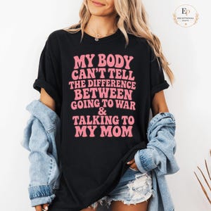 Peut inclure: T-shirt noir avec l'inscription rose "MY BODY CAN'T TELL THE DIFFERENCE BETWEEN GOING TO WAR & TALKING TO MY MOM". Le t-shirt est porté avec une veste en jean et un short.