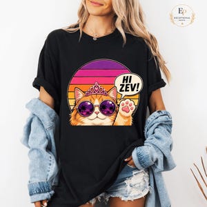 Könnte beinhalten: Schwarzes T-Shirt mit einer Cartoon-Katze, die eine Sonnenbrille und eine Tiara trägt, vor einem Retro-Sonnenuntergangsdesign. Die Katze winkt mit einer Pfote und eine Sprechblase sagt "Hi Zev!"