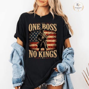 Peut inclure: T-shirt noir avec un drapeau américain vieilli, une silhouette de soldat et le texte "ONE BOSS NO KINGS". Le graphisme utilise des tons marron, rouge et bleu.