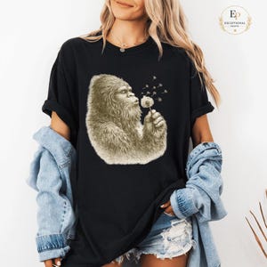 Peut inclure: T-shirt noir avec une illustration sépia d'un Bigfoot soufflant sur un pissenlit. Le Bigfoot est représenté avec des poils détaillés et une expression contemplative. Les graines de pissenlit s'envolent de la fleur.