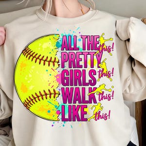 May include: A cream-coloured sweatshirt with a large softball graphic. The text reads "ALL THE PRETTY GIRLS WALK LIKE THIS!" in pink and yellow. The softball is yellow with red stitching and colourful paint splatters.