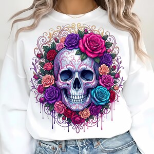 May include: A white long-sleeve sweatshirt featuring a central graphic of a human skull adorned with intricate pink filigree patterns. The skull is surrounded by a vibrant wreath of roses in shades of red, pink, purple, and blue with paint drips.