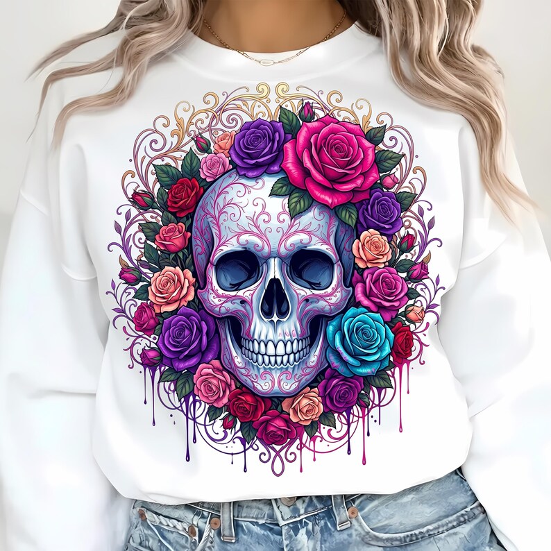 May include: A white long-sleeve sweatshirt featuring a central graphic of a human skull adorned with intricate pink filigree patterns. The skull is surrounded by a vibrant wreath of roses in shades of red, pink, purple, and blue with paint drips.