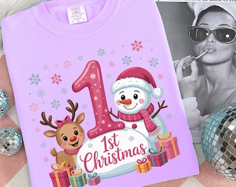 Può includere: Una maglietta lilla a maniche lunghe con un design natalizio. Il design presenta un grande numero 1, un pupazzo di neve con cappello e sciarpa, una renna e la scritta "1st Christmas". Regali e fiocchi di neve completano il design.
