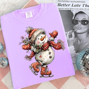 May include: A light purple sweatshirt featuring a whimsical snowman illustration. The snowman wears a red hat, scarf, and mittens, and is ice skating. The design includes a carrot nose, a sprig of holly, and the text "IMA CONCE".