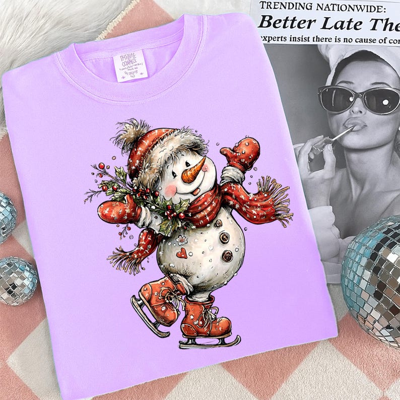 May include: A light purple sweatshirt featuring a whimsical snowman illustration. The snowman wears a red hat, scarf, and mittens, and is ice skating. The design includes a carrot nose, a sprig of holly, and the text "IMA CONCE".