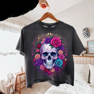 May include: A charcoal gray t-shirt featuring a central graphic of a human skull surrounded by a vibrant arrangement of roses in shades of pink, purple, red, and blue, accented with intricate gold filigree swirls and subtle paint drip effects.