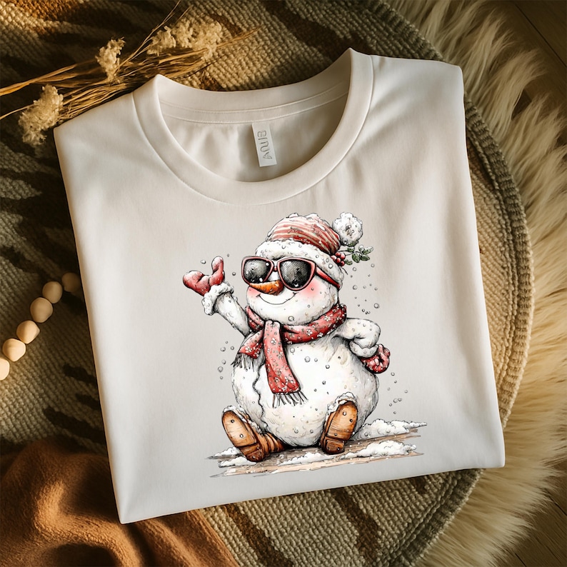 May include: Off-white t-shirt featuring a cartoon snowman wearing sunglasses, a red and white striped hat, and a matching scarf. The snowman is sitting in the snow and wearing brown boots. A winter-themed graphic tee.