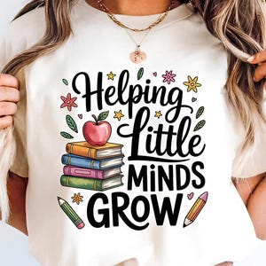 May include: Cream-colored t-shirt featuring the text "Helping Little Minds Grow" in black script. The design includes a stack of books, a red apple, pencils, and floral accents in various colors. A teacher-themed graphic.