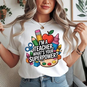 May include: White t-shirt with the text "I'M A TEACHER, WHAT'S YOUR SUPERPOWER?" surrounded by school supplies like a red apple, pencils, crayons, and scissors. The shirt is a crew neck style.