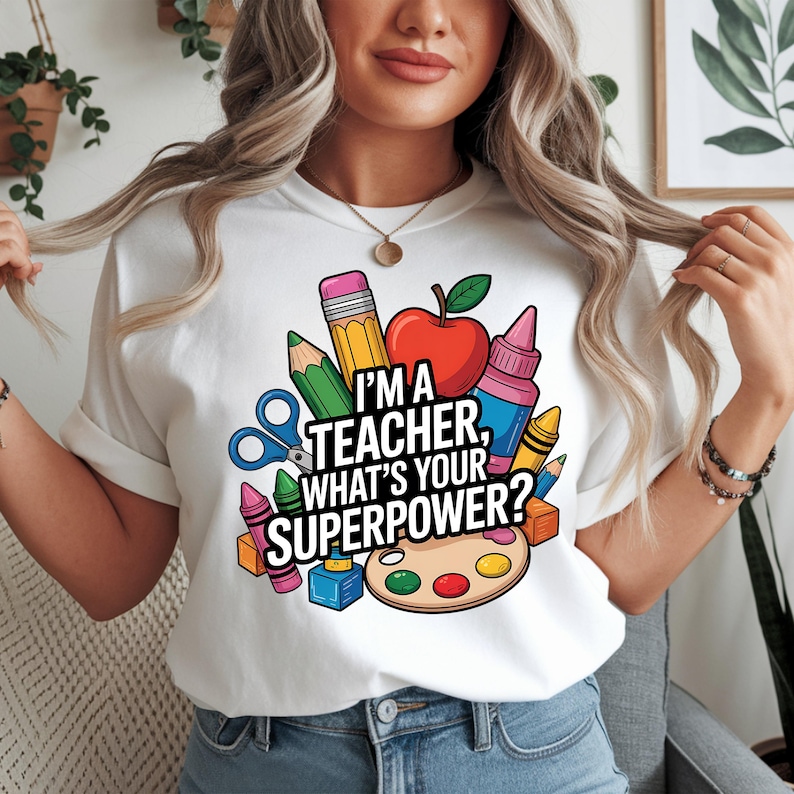 May include: White t-shirt with the text "I'M A TEACHER, WHAT'S YOUR SUPERPOWER?" surrounded by school supplies like a red apple, pencils, crayons, and scissors. The shirt is a crew neck style.