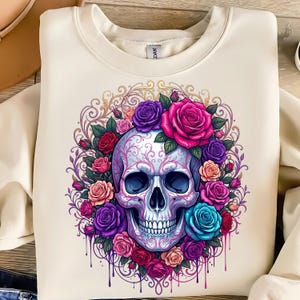 May include: A cream-colored crewneck sweatshirt featuring a central graphic of a human skull adorned with intricate filigree patterns, surrounded by a vibrant wreath of multi-colored roses in shades of pink, purple, red, and blue with paint drips.
