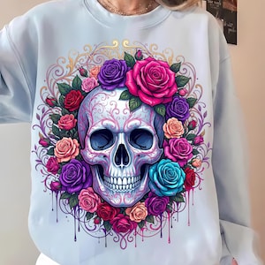 May include: A light blue crewneck sweatshirt featuring a central graphic of a human skull adorned with intricate pink filigree patterns. The skull is surrounded by a vibrant wreath of multi-colored roses in shades of red, pink, purple, and teal.