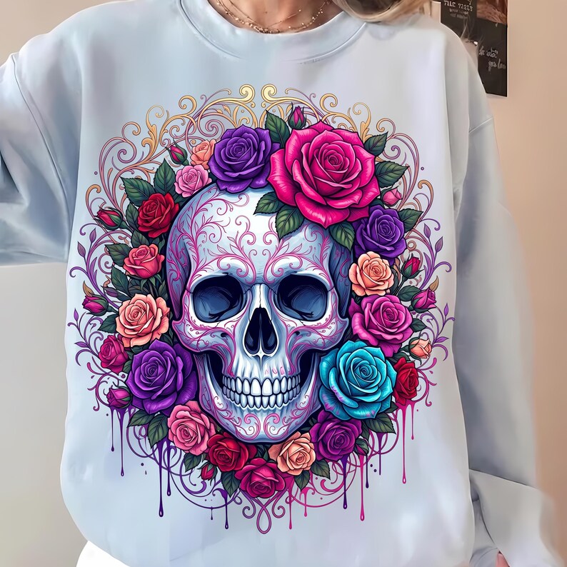 May include: A light blue crewneck sweatshirt featuring a central graphic of a human skull adorned with intricate pink filigree patterns. The skull is surrounded by a vibrant wreath of multi-colored roses in shades of red, pink, purple, and teal.