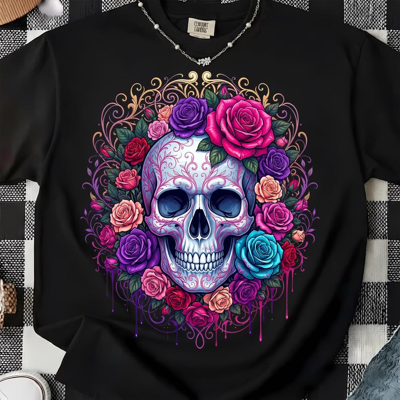 May include: A black t-shirt featuring a central graphic of a white skull adorned with intricate pink filigree patterns. The skull is surrounded by a vibrant arrangement of roses in shades of red, pink, purple, and blue, set against a gold filigree frame.