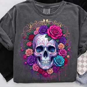 May include: A charcoal gray t-shirt featuring a central graphic of a human skull adorned with intricate pink filigree patterns. The skull is surrounded by a vibrant arrangement of roses in shades of red, pink, purple, and blue with green leaves.