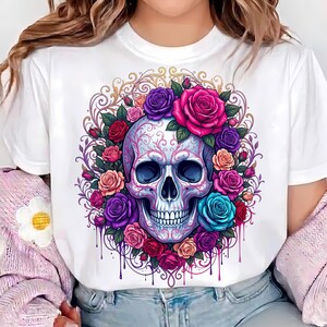May include: A white t-shirt featuring a central graphic of a human skull adorned with intricate filigree patterns, surrounded by a vibrant wreath of multi-colored roses in shades of pink, purple, red, orange, and blue, with dripping paint accents.