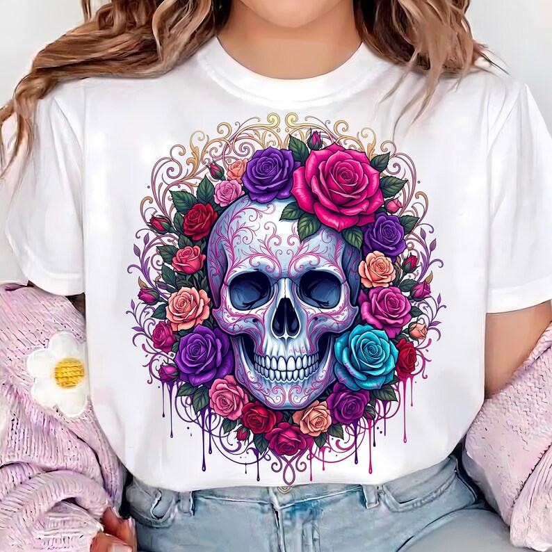 May include: A white t-shirt featuring a central graphic of a human skull adorned with intricate filigree patterns, surrounded by a vibrant wreath of multi-colored roses in shades of pink, purple, red, orange, and blue, with dripping paint accents.