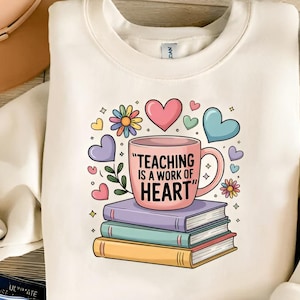 May include: Cream-colored sweatshirt featuring a colorful graphic. The graphic includes a pink mug with the text "TEACHING IS A WORK OF HEART" atop a stack of books, surrounded by hearts and flowers. A hat and a small plant are visible.