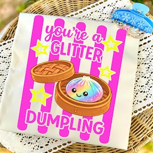 May include: A cream-colored t-shirt with a graphic of a rainbow dumpling in a bamboo steamer. The text reads "You're a Glitter Dumpling" in pink, with yellow star accents and pink vertical stripes.