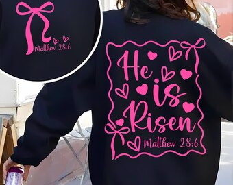 Può includere: Felpa nera con grafica rosa. La parte anteriore presenta un fiocco e la scritta "Matthew 28:6". Il retro riporta la scritta "He is Risen" con cuori e fiocchi, e la scritta "Matthew 28:6".