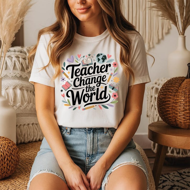 May include: A white t-shirt with the text "Teacher Change the World" in a black, cursive font, surrounded by colorful illustrations of pencils, books, flowers, and a globe. The shirt is a crew neck style.