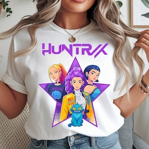 Puede incluir: Camiseta blanca con un diseño de estrella púrpura que presenta a tres mujeres de dibujos animados y un gato azul. La camiseta muestra la palabra "HUNTR/X" en púrpura. Las mujeres tienen diferentes colores de cabello y usan varios atuendos. El gato tiene ojos grandes.