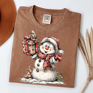 May include: A brown t-shirt featuring a cheerful snowman illustration. The snowman wears a red and white hat and scarf, holding a gift box with a red ribbon. The design includes a small snowman and snow details, perfect for the holiday season.