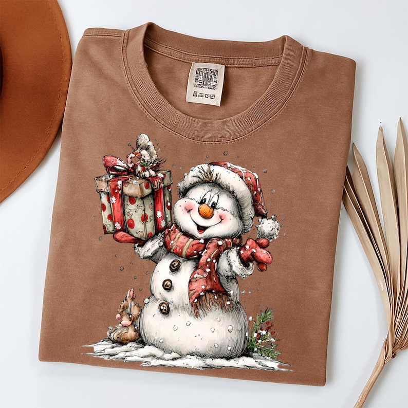 May include: A brown t-shirt featuring a cheerful snowman illustration. The snowman wears a red and white hat and scarf, holding a gift box with a red ribbon. The design includes a small snowman and snow details, perfect for the holiday season.