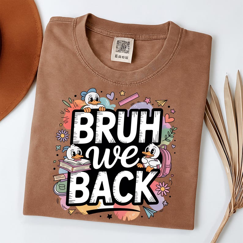 May include: A brown t-shirt with the text "BRUH we BACK" in white, surrounded by cartoon ducks, flowers, and school-themed graphics. The shirt is made of a soft material and has a crew neck.