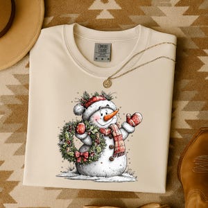 May include: Cream-colored sweatshirt featuring a cheerful snowman illustration. The snowman wears a red and white hat, scarf, and mittens, holding a wreath and a mug. The sweatshirt has the Comfort Colors logo.