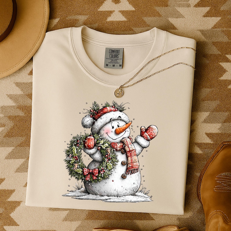 May include: Cream-colored sweatshirt featuring a cheerful snowman illustration. The snowman wears a red and white hat, scarf, and mittens, holding a wreath and a mug. The sweatshirt has the Comfort Colors logo.