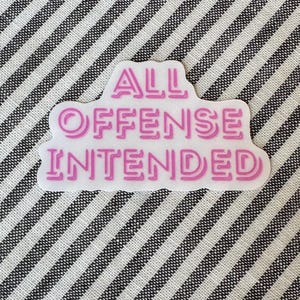May include: A white sticker with the text "ALL OFFENSE INTENDED" in pink, outlined in a darker pink. The sticker is on a striped black and white fabric background.
