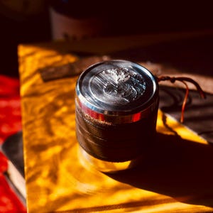 May include: A cylindrical grinder with a silver top and a dark brown leather-like body. The top has a detailed design. A small leather strap is attached. The grinder sits on a yellow surface, with a red surface in the background.